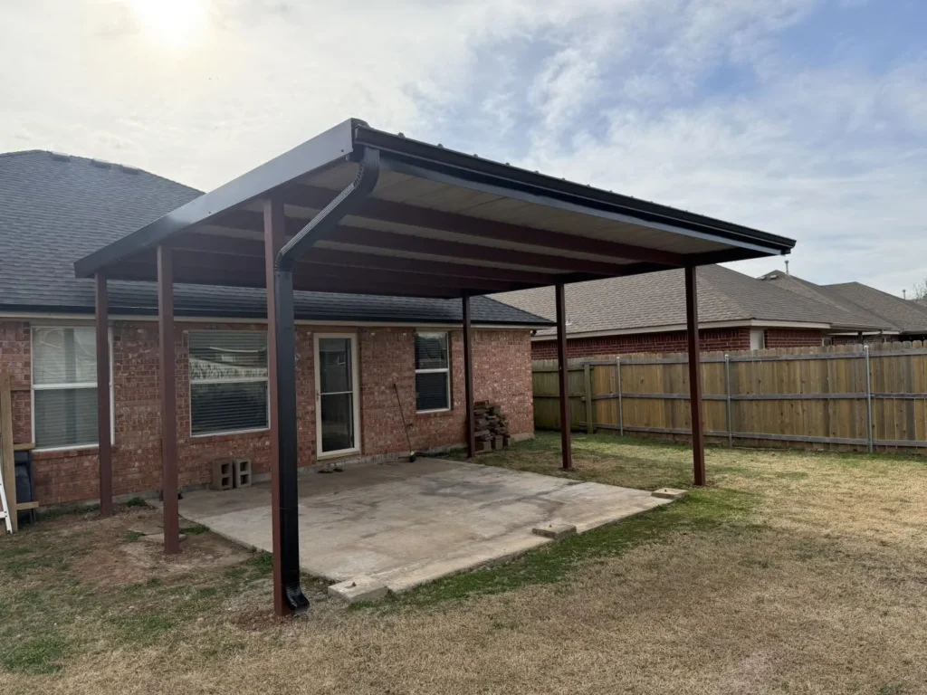 A metal patio cover attached to the side of a brick house over a concrete slab.