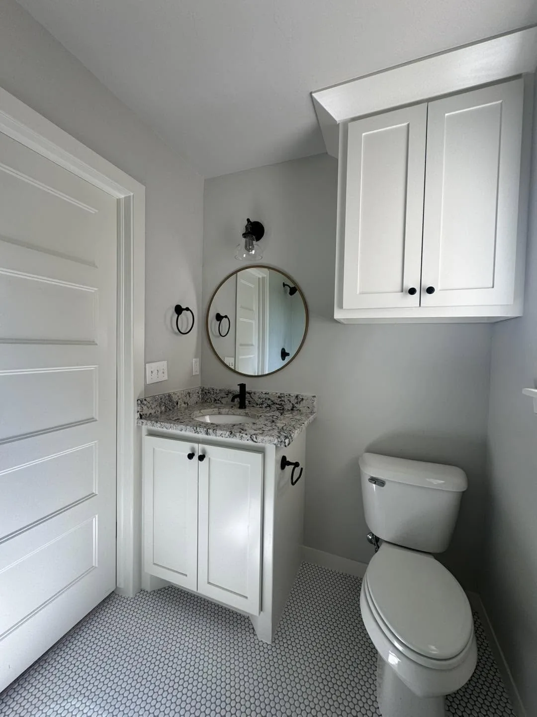 A bathroom with a white vanity, a speckled granite countertop, a circular mirror, wall-mounted storage cabinets, and a toilet on a penny-tile floor.