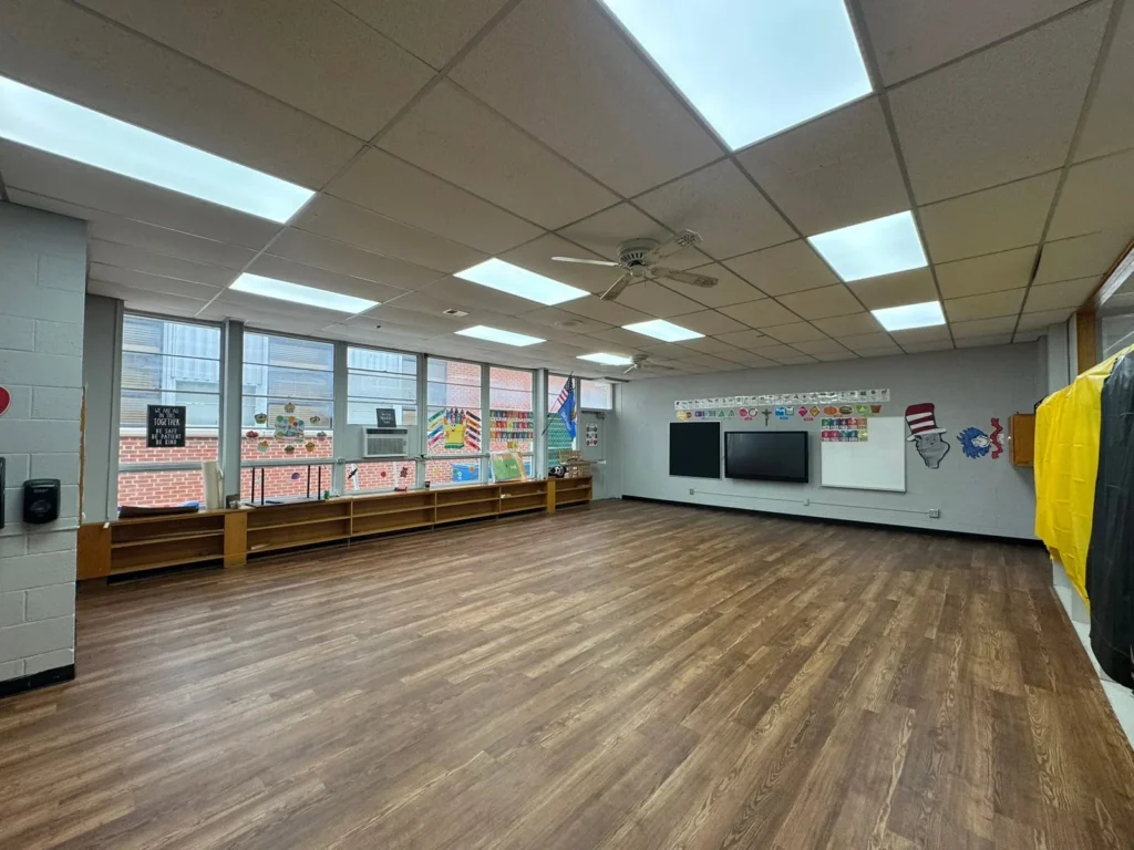 An empty classroom with wood-look flooring, large windows, and a wall featuring a television screen and decorative cutouts.