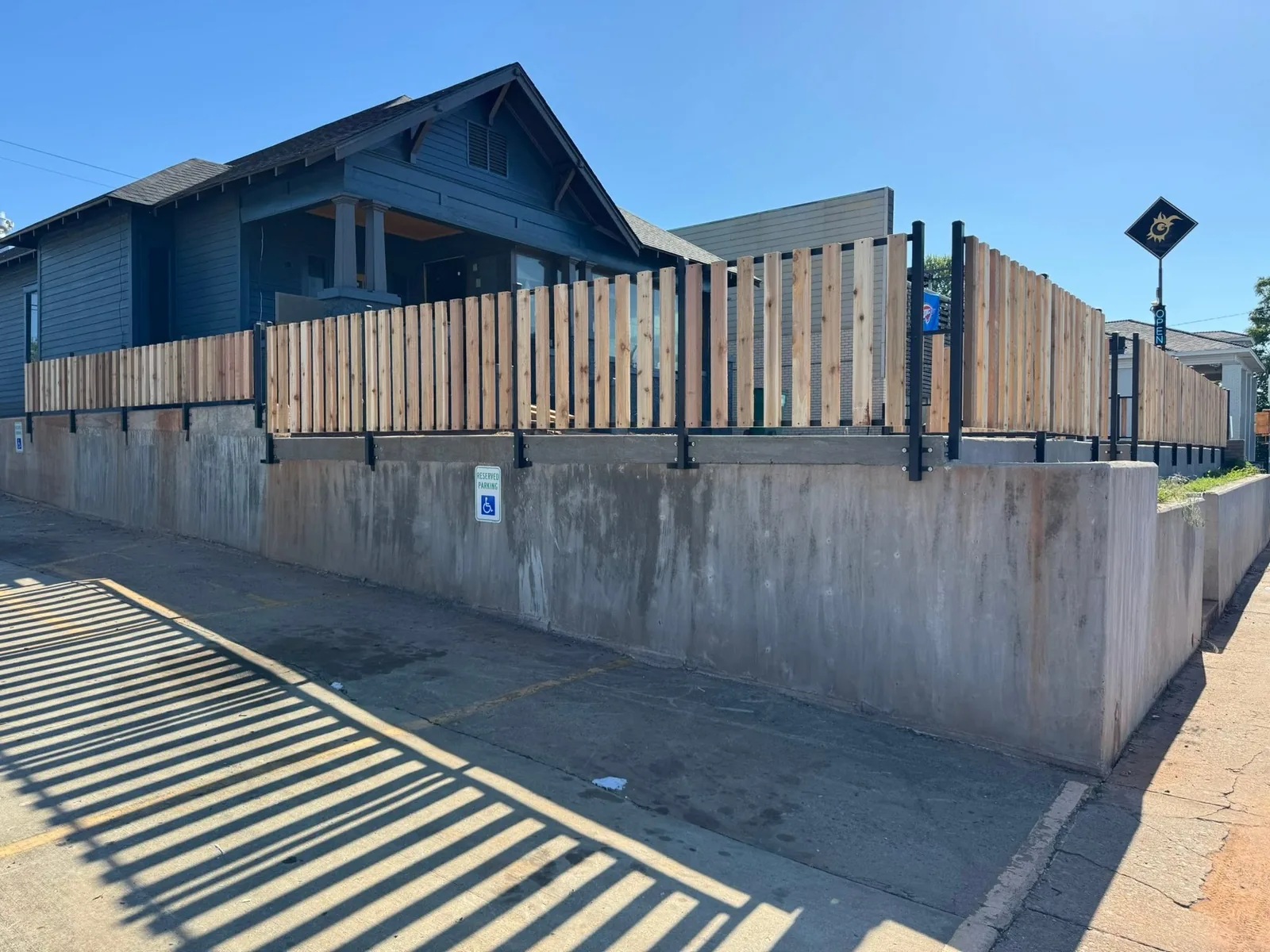 A blue house behind a tall concrete retaining wall topped with a new wooden slat fence and a small handicap parking sign.