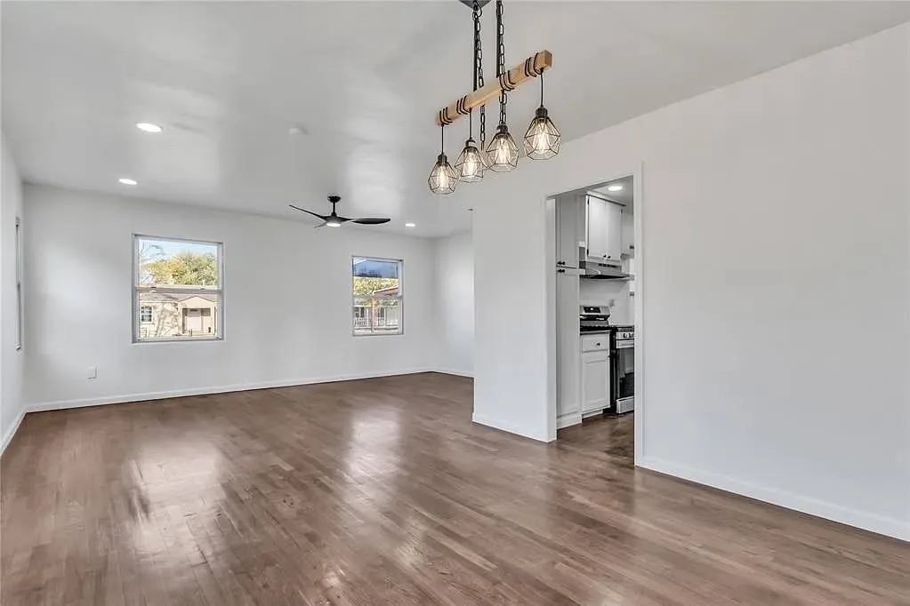 An open living area with hardwood floors, a hanging chandelier, and a view into a kitchen.