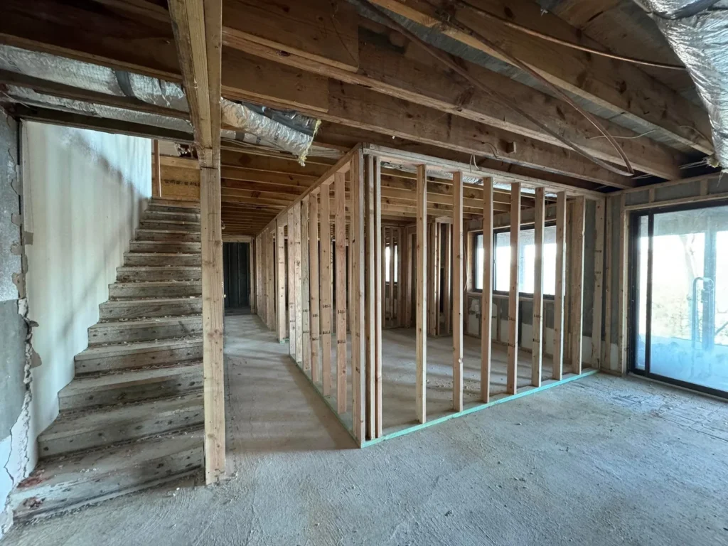 Unfinished interior space under construction with exposed wooden wall framing, an open staircase, and concrete flooring.