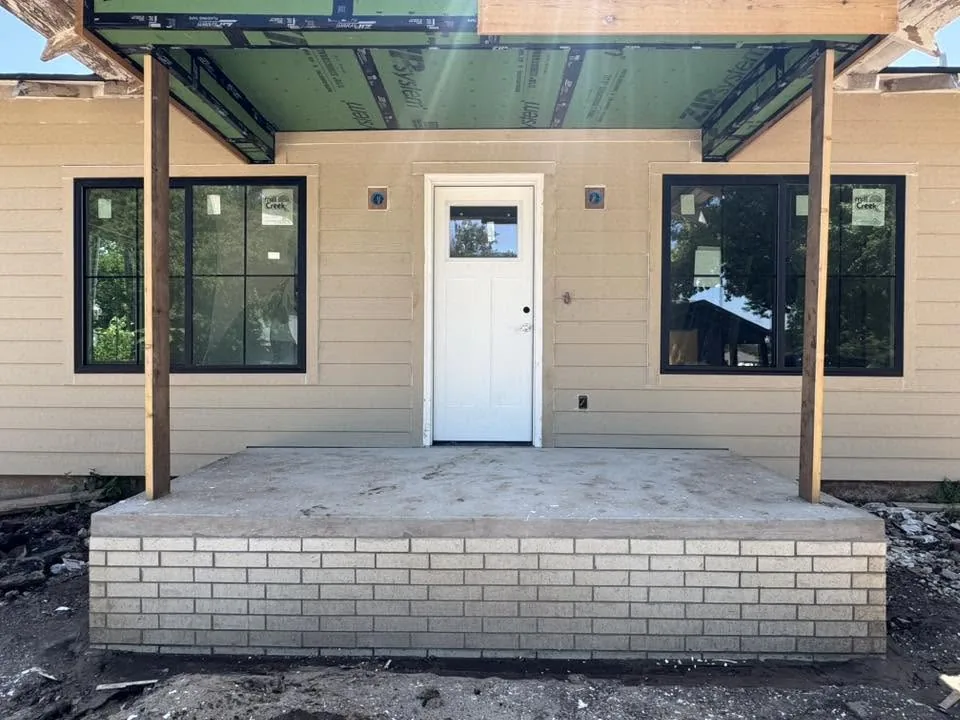 A house exterior under construction featuring a concrete porch, a white front door, and two black-framed windows.