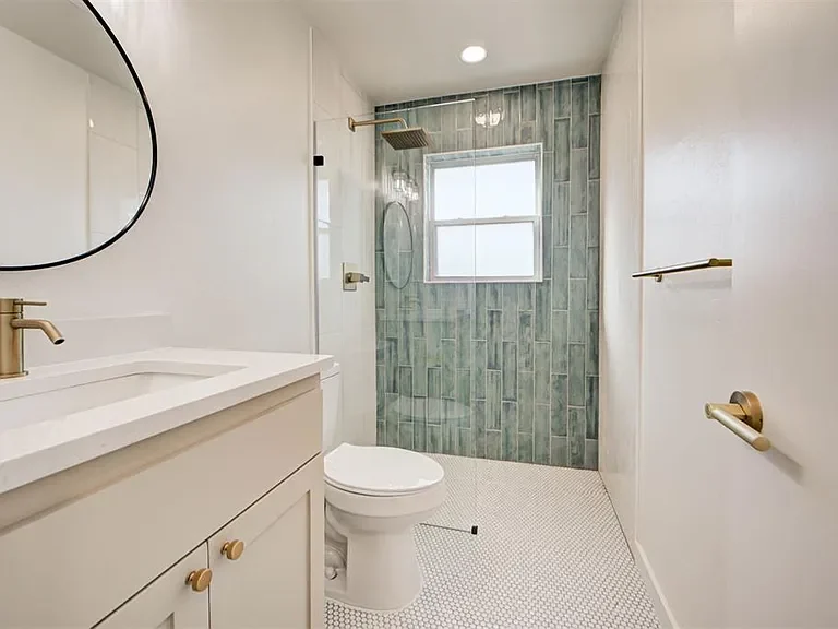 Bathroom with a white vanity, a round mirror, and a walk-in shower with green subway tile and white hexagonal floor tiles.