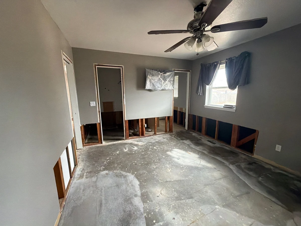 A room undergoing renovation with exposed wall studs at the floor level, bare concrete floors, and a covered TV mounted on the wall.
